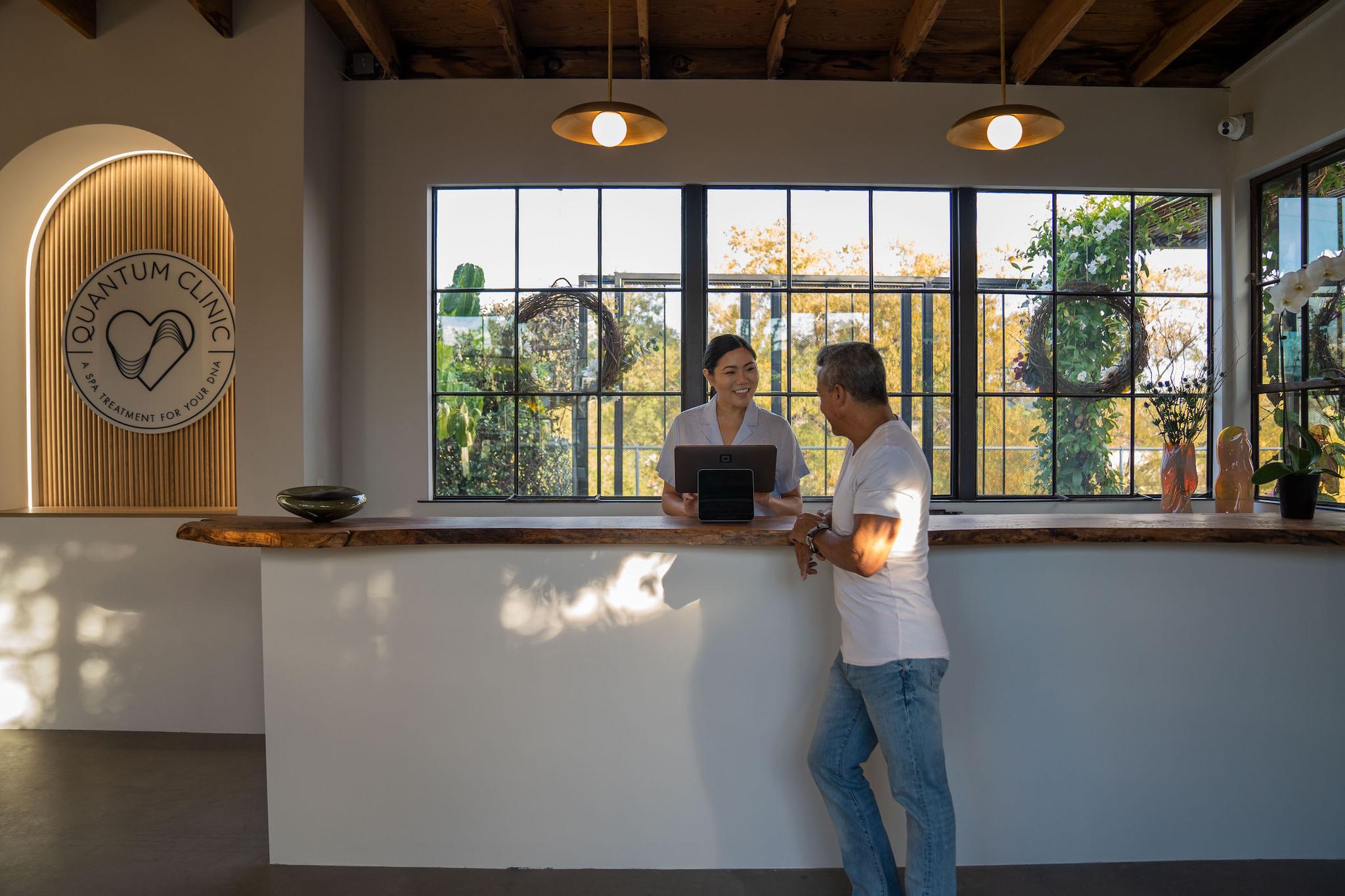 Front desk at Quantum Clinic with a receptionist warmly welcoming a guest in a bright, nature-filled wellness center